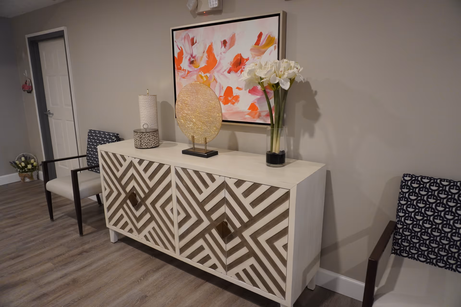 A hallway or waiting area with a decorative white and brown patterned cabinet against a gray wall. On top of the cabinet are a vase with white flowers, a round gold decorative piece, and a candle holder. Above the cabinet hangs a colorful abstract painting with pink, orange, and white tones. Two chairs with dark wooden arms and patterned cushions are placed on either side of the cabinet. The floor is wood laminate, and there is a closed white door in the background.