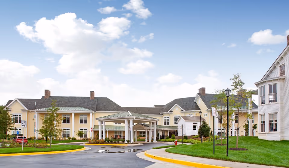 Exterior front view of a large yellow senior living facility with a covered drop-off entrance, driveway, and landscaped grounds.
