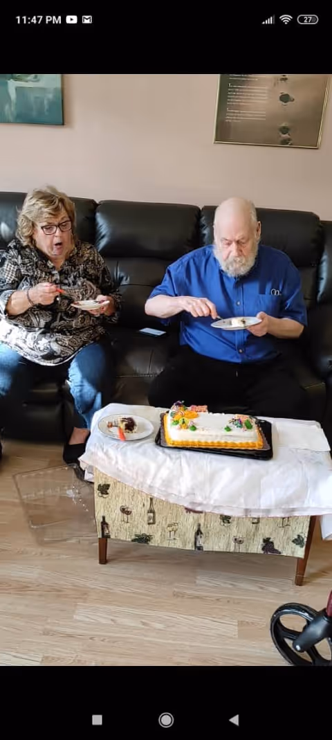Two elderly individuals sitting on a black leather couch eating cake from plates. A decorated cake is placed on a small table covered with a white cloth in front of them. The room has light-colored wooden flooring and framed pictures on the wall behind them.