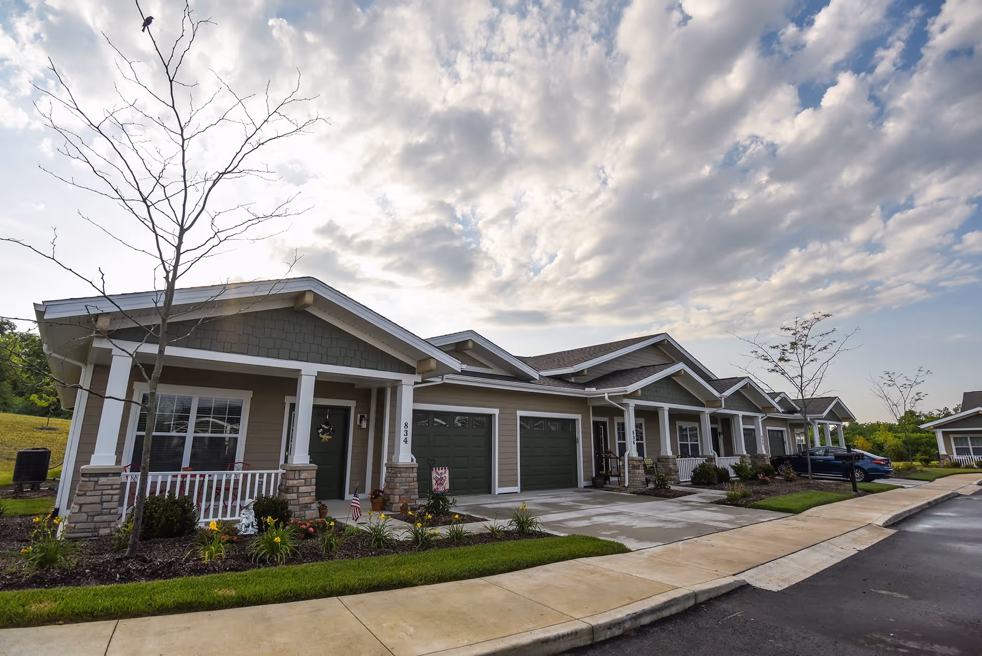 Row of single-story residential units with garages and small front porches, surrounded by landscaped greenery and young trees under a partly cloudy sky.