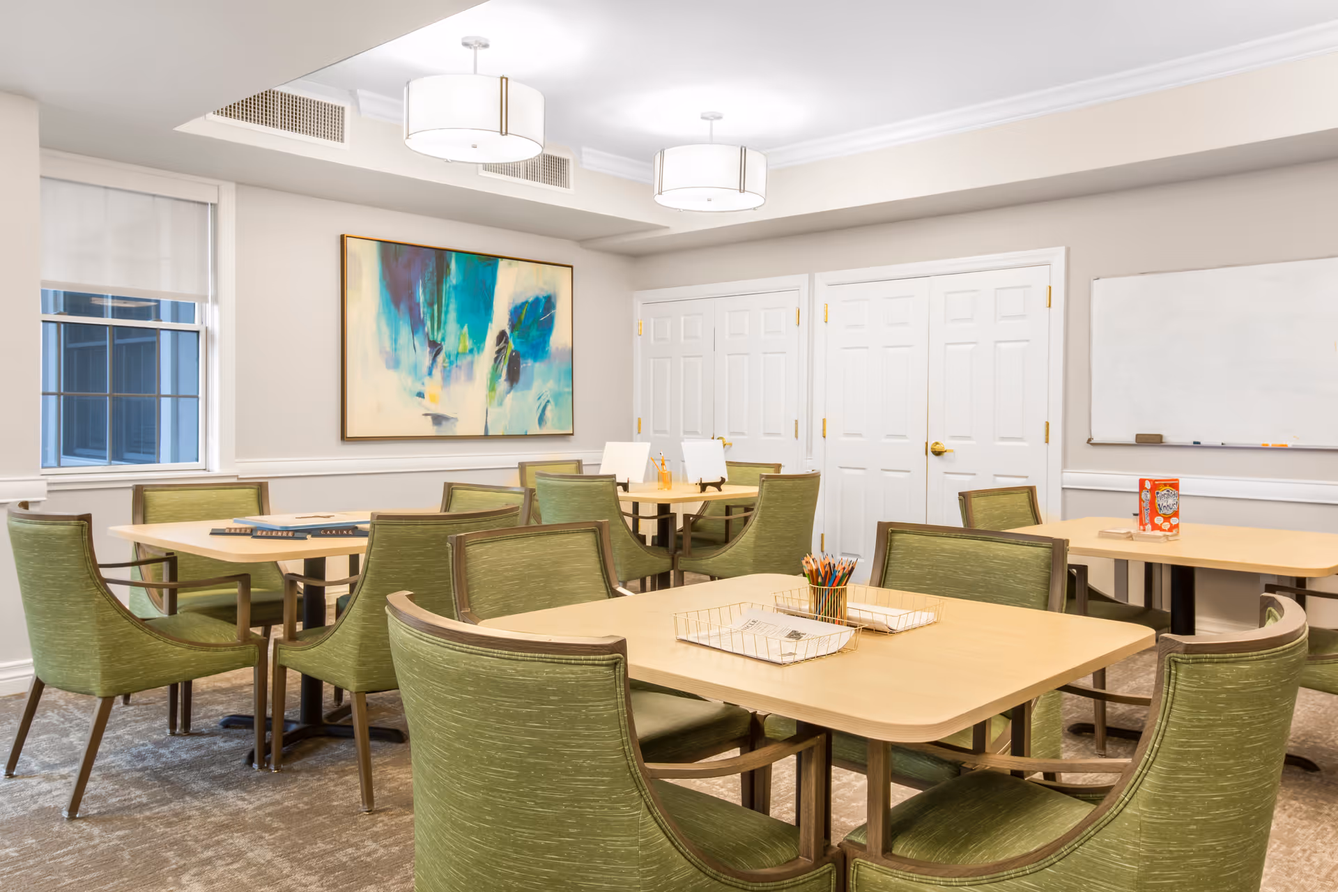 A bright and clean activity or meeting room with multiple light wood tables surrounded by green upholstered chairs. The room features a large abstract painting on one wall, a whiteboard on another, and two modern ceiling light fixtures. There are windows letting in natural light and several closed white doors.