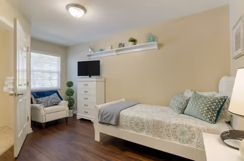 A cozy bedroom with a single bed featuring patterned bedding and multiple pillows. There is a white dresser with a small flat-screen TV on top, a white armchair with a blue throw blanket, and a small green plant beside it. The room has wooden flooring, beige walls, a window with blinds, and a white door partially open.