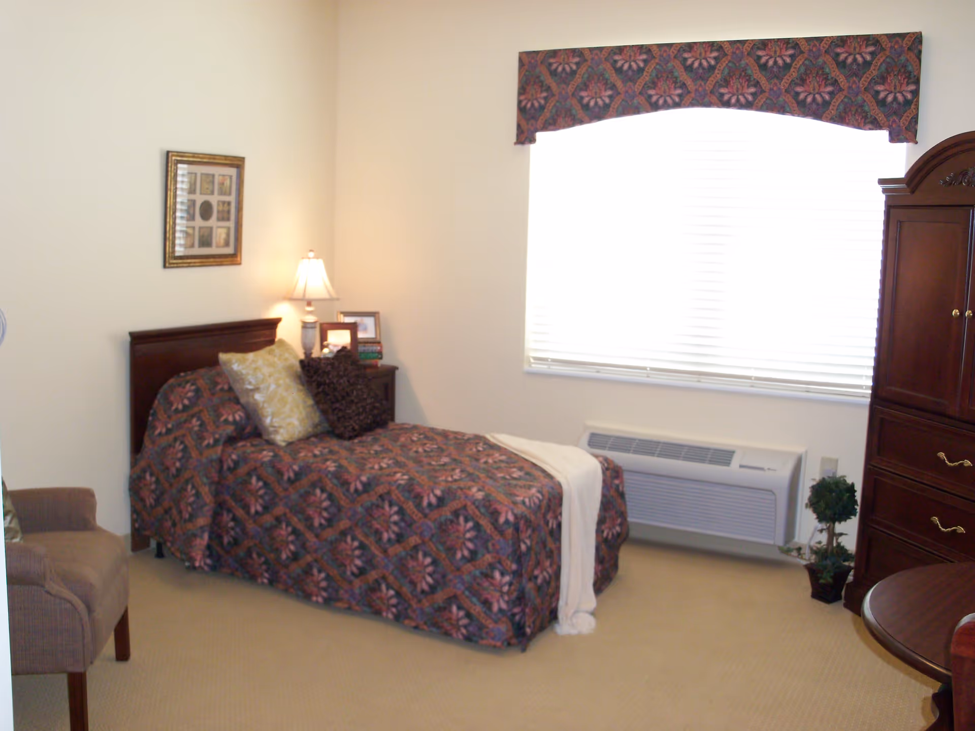 Small furnished bedroom with a single bed, bedside table and lamp, large window with blinds and a wooden armoire.