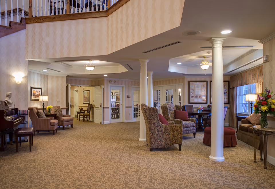 Spacious furnished lounge in an assisted living facility with upholstered chairs, columns, a piano, and decorative lighting.