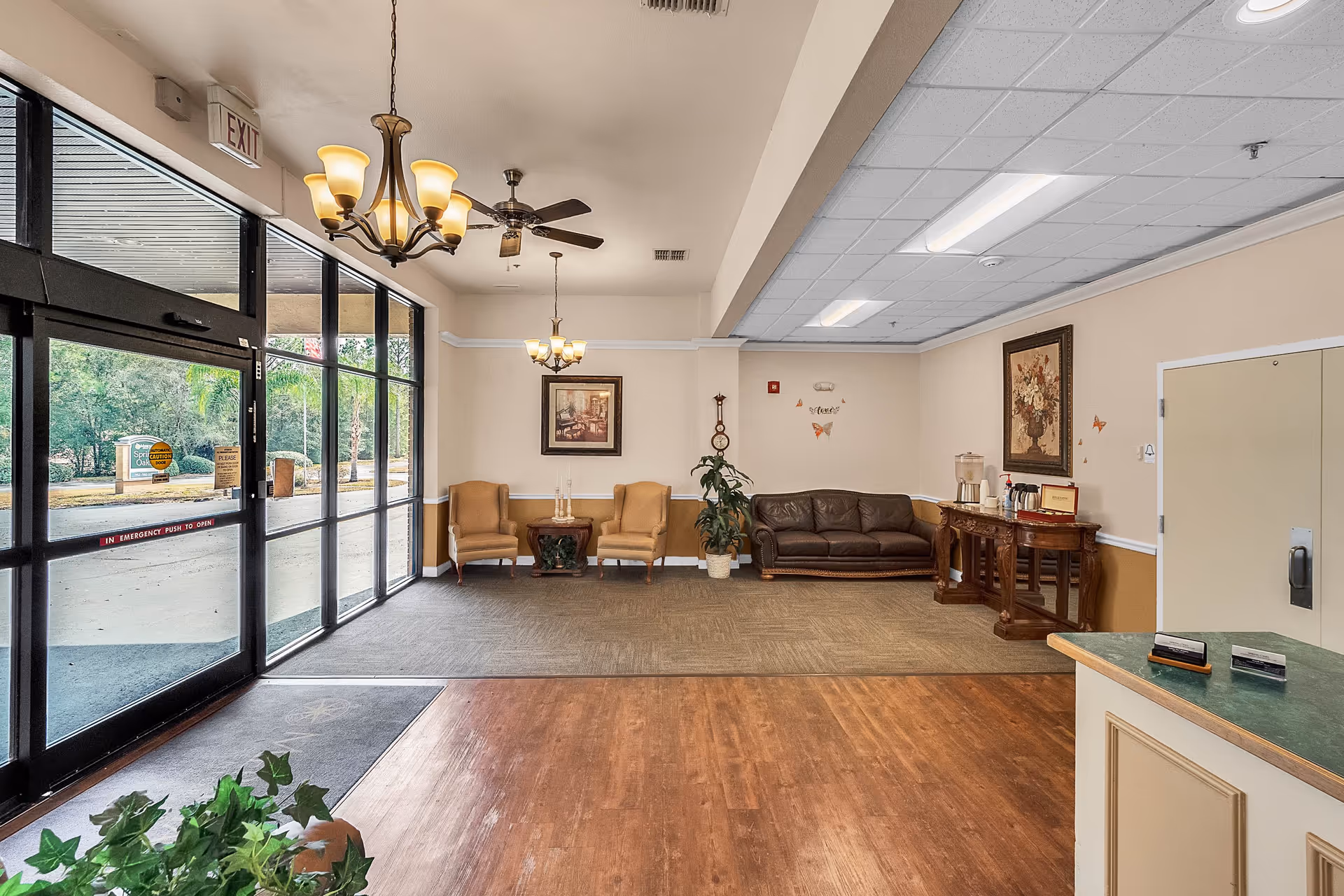 Interior view of a senior living facility lobby with large glass entrance doors on the left, two beige armchairs and a small table against the back wall, a brown leather sofa, a wooden console table with a water dispenser and coffee setup, framed artwork on the walls, ceiling lights, and a ceiling fan.