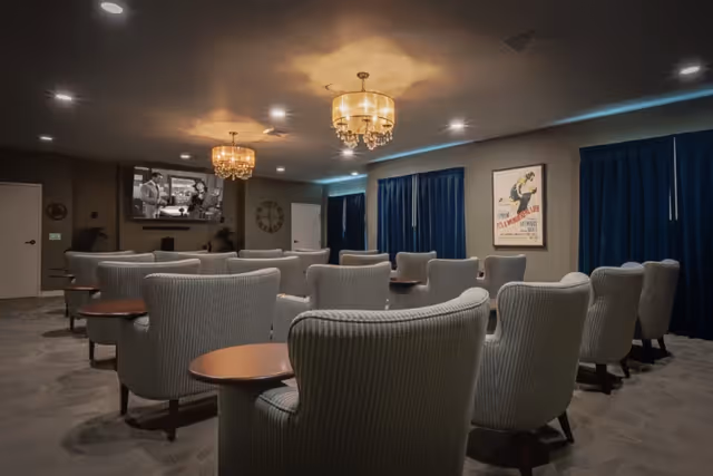 A cozy theater room with multiple rows of comfortable armchairs facing a large wall-mounted TV screen. The room features two elegant chandeliers, dark blue curtains covering the windows, and a framed vintage movie poster on the wall.
