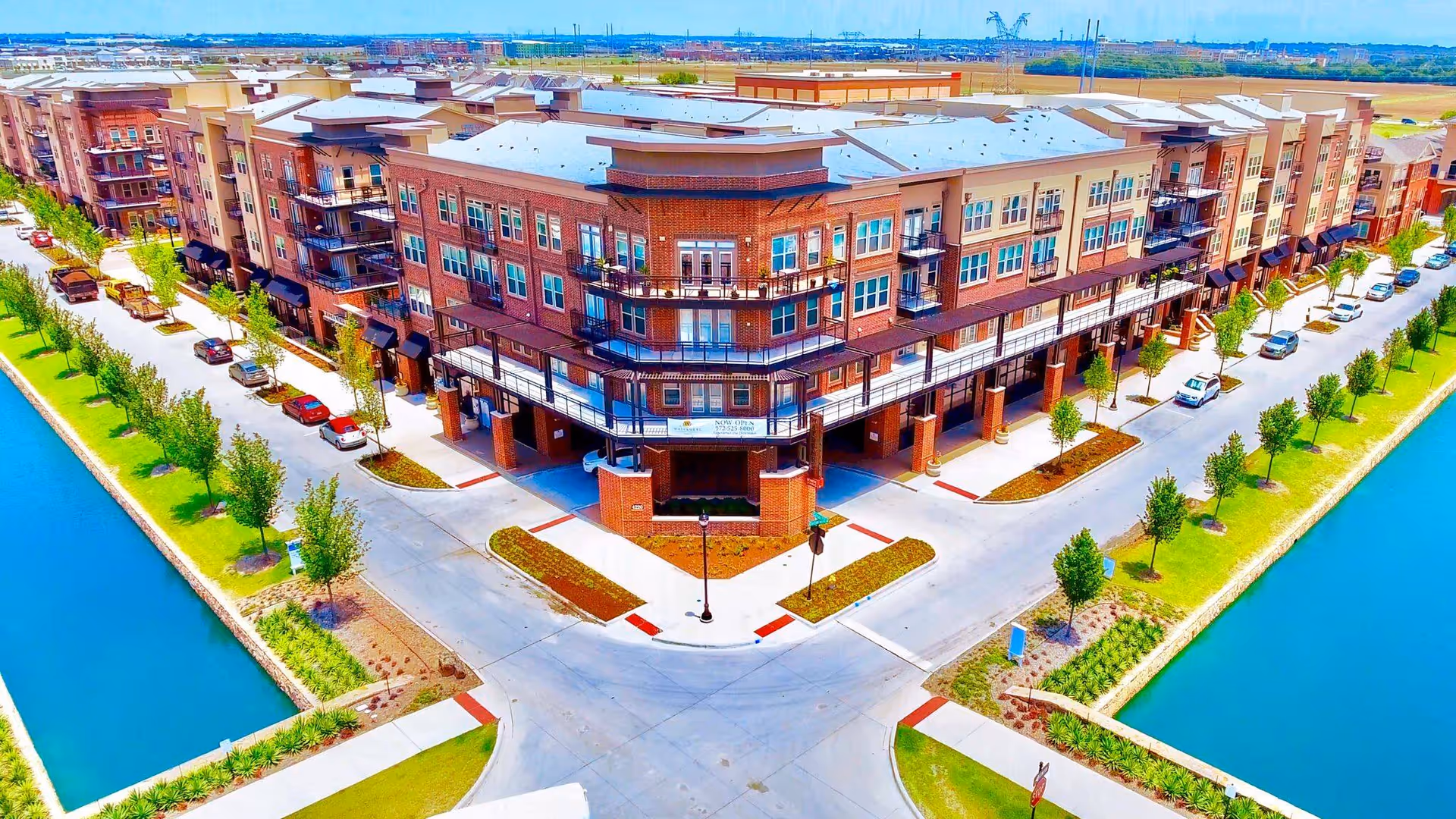 Aerial view of a large, modern multi-story residential building complex with brick and beige exterior, surrounded by roads, parked cars, and landscaped green areas with trees. The complex is bordered by a water canal on two sides.