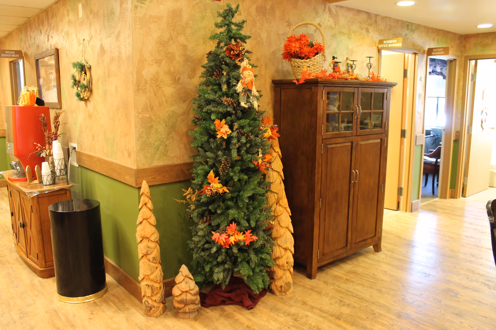 Interior hallway corner of a senior living facility decorated with a Christmas tree adorned with autumn leaves and pine cones, two wooden carved tree sculptures, a wooden cabinet topped with a basket of orange flowers and small decorative items, and a beverage station with cups and a red dispenser. Doors labeled 'Business Office Manager' and 'Dietary Manager' are visible in the background.