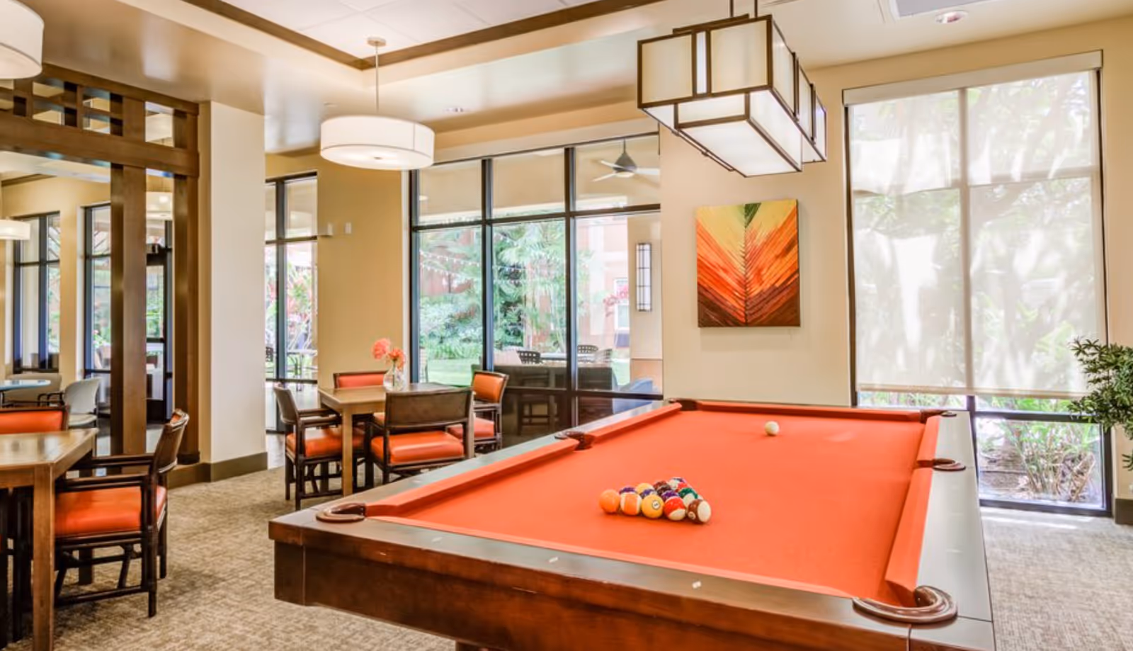 A bright and inviting game room featuring a red felt pool table with billiard balls arranged for a game. The room has large windows letting in natural light, modern pendant lights, and a seating area with tables and chairs upholstered in orange and brown. A colorful abstract painting hangs on the wall, and greenery is visible outside the windows.