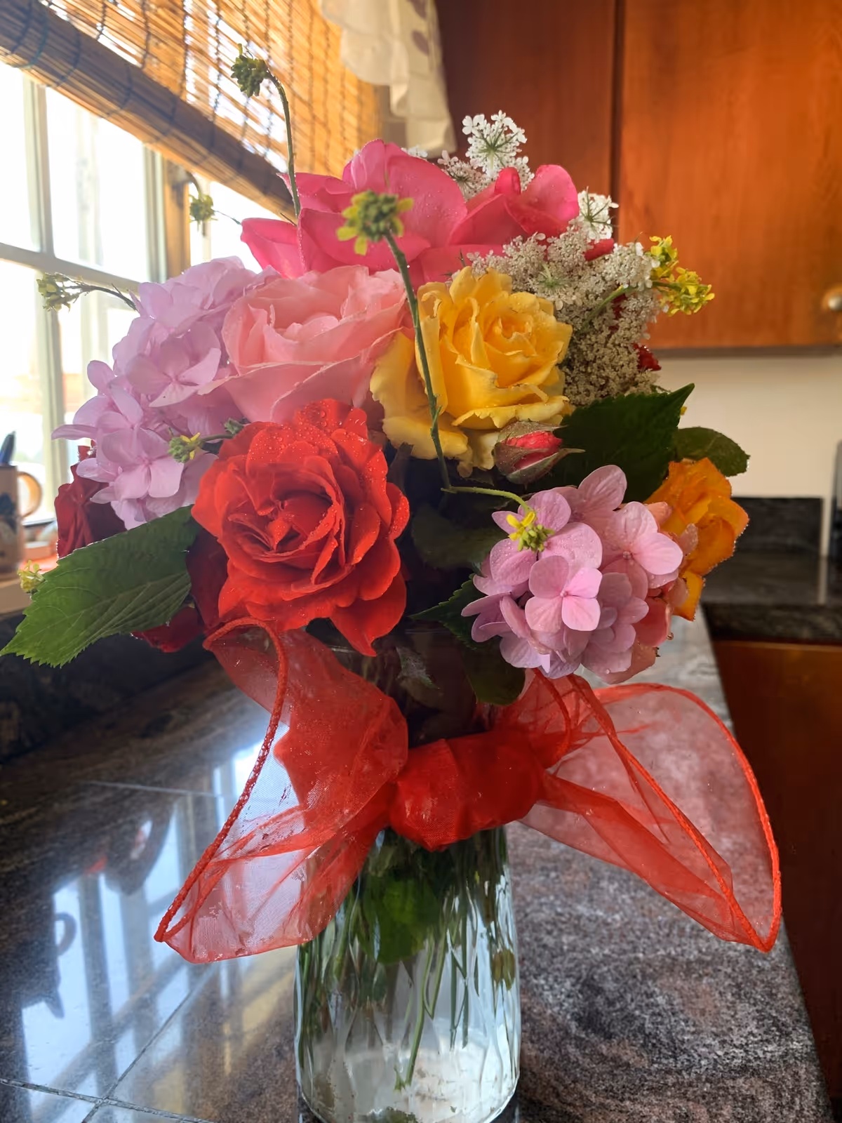 A colorful bouquet of flowers including red, pink, yellow, and purple blooms arranged in a clear glass vase with a large red ribbon tied around it, placed on a kitchen countertop with wooden cabinets and a window with bamboo blinds in the background.