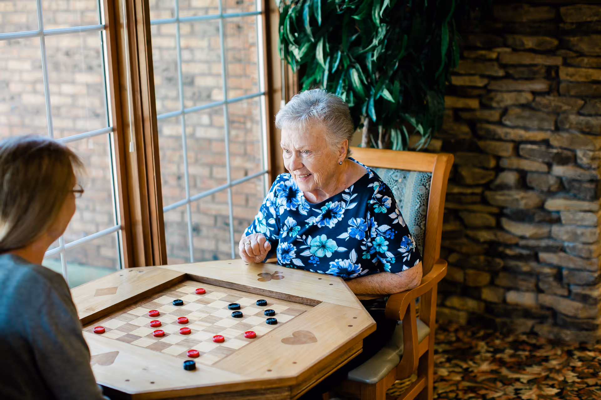 Two elderly women sitting at a wooden table with a built-in checkerboard, playing a game of checkers in a room with large windows, a stone wall, and a large green plant in the background.