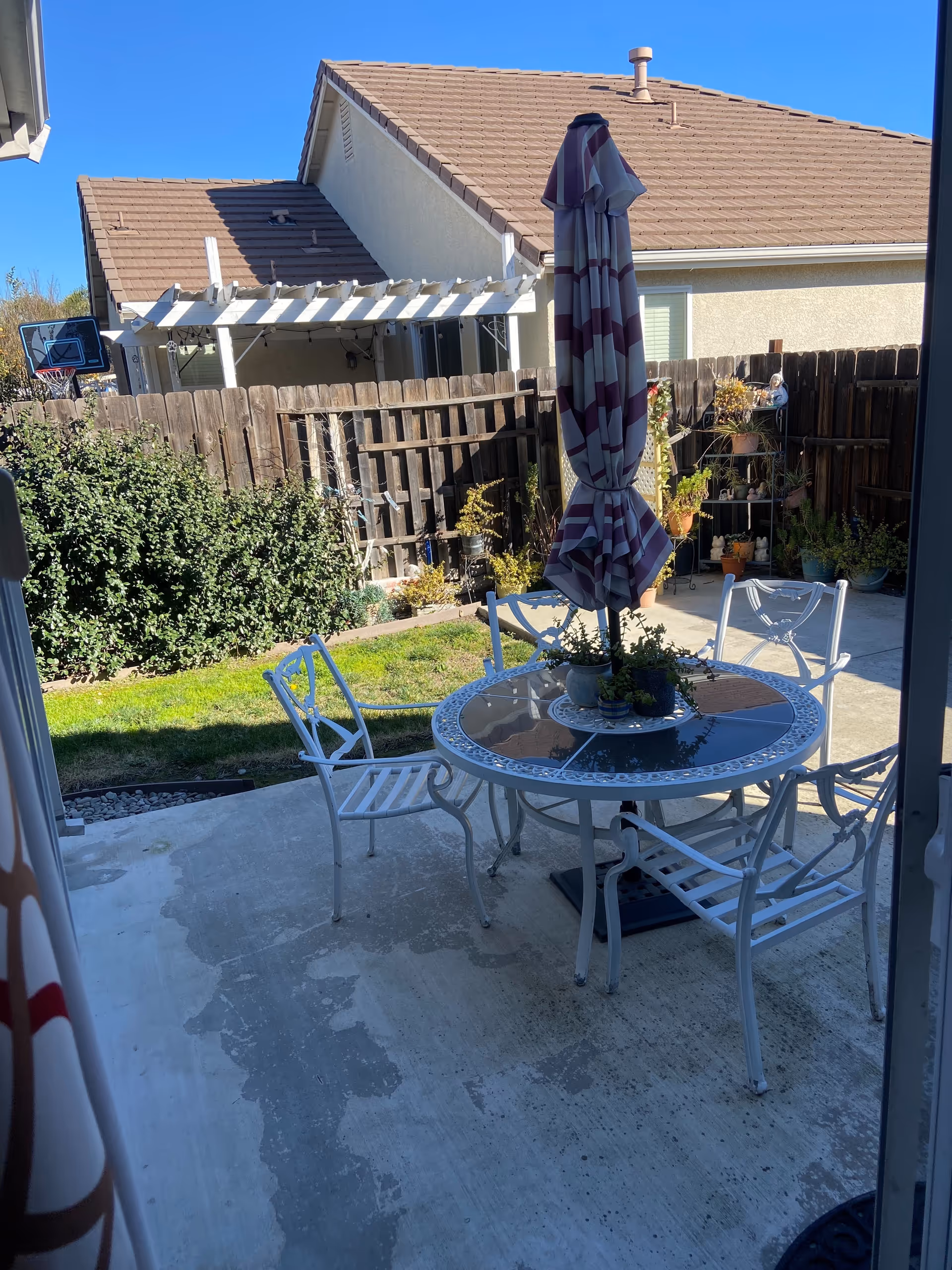 Outdoor patio area with a round glass-top table surrounded by four white metal chairs. A closed striped umbrella is positioned in the center of the table. The patio is concrete and adjacent to a grassy area with plants and a wooden fence. In the background, there are neighboring houses with beige walls and brown roofs.