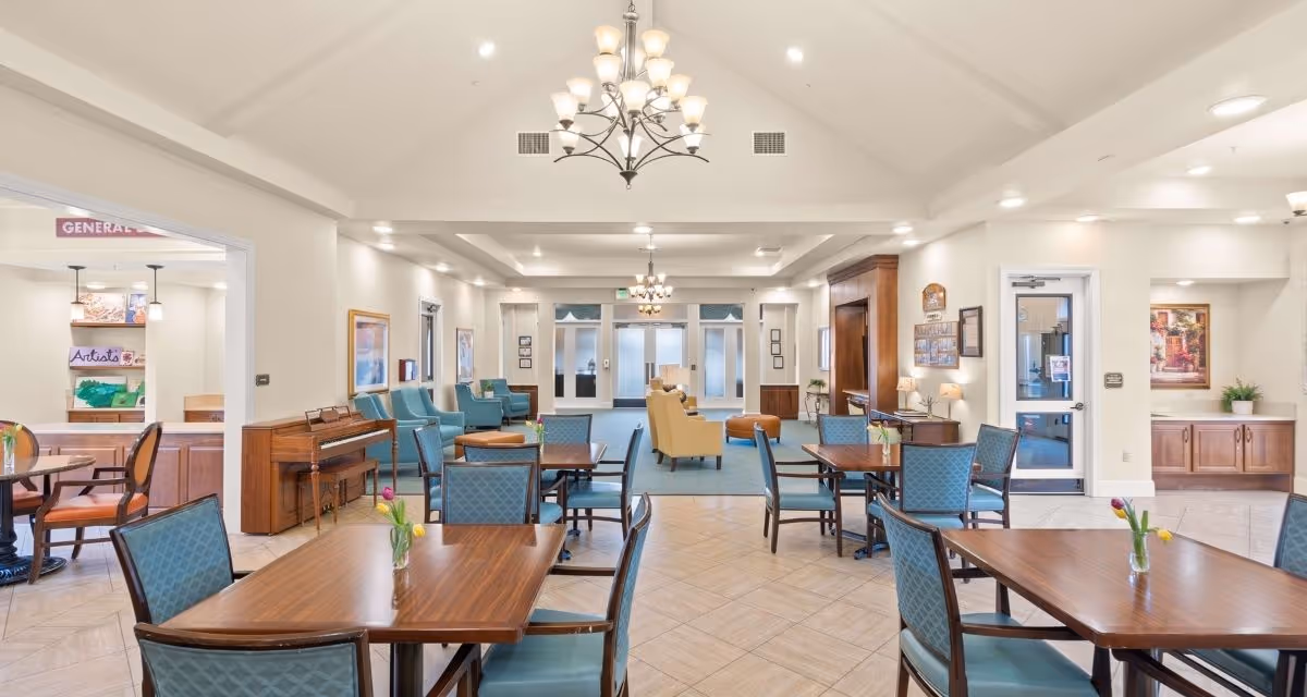 A spacious and well-lit common area in a senior living facility featuring multiple wooden tables with blue cushioned chairs. There are flower vases on the tables, a piano against the left wall, and comfortable armchairs arranged near the back. The room has high ceilings with chandeliers and recessed lighting. To the left, there is an entrance to an art room with a sign that reads 'Artists'. The walls are decorated with framed artwork and there is a door leading outside on the right.