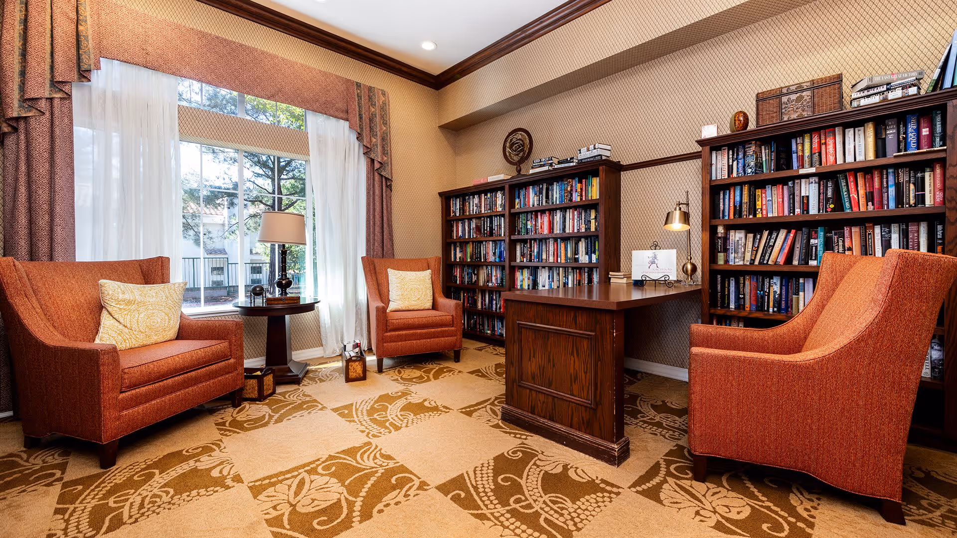 A cozy reading room with three orange upholstered armchairs arranged around a wooden desk. Behind the desk are two tall bookshelves filled with books. A large window with sheer white curtains and patterned drapes lets in natural light, and a small round table with a lamp sits between two of the chairs. The carpet has a decorative floral pattern in beige and brown tones.