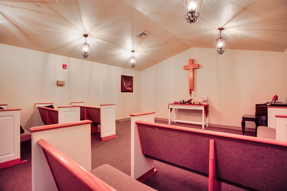 Interior of a small chapel or prayer room with wooden pews arranged facing a wall with a large wooden cross mounted above a small table. The table holds a Bible and other religious items. The room is softly lit by hanging lantern-style light fixtures, and there is a framed religious painting on one wall.