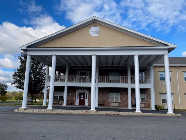 Front exterior view of a two-story building with a large covered entrance supported by six white columns. The building has beige and brick walls, multiple windows, and a peaked roof with a vent. The sky is partly cloudy.