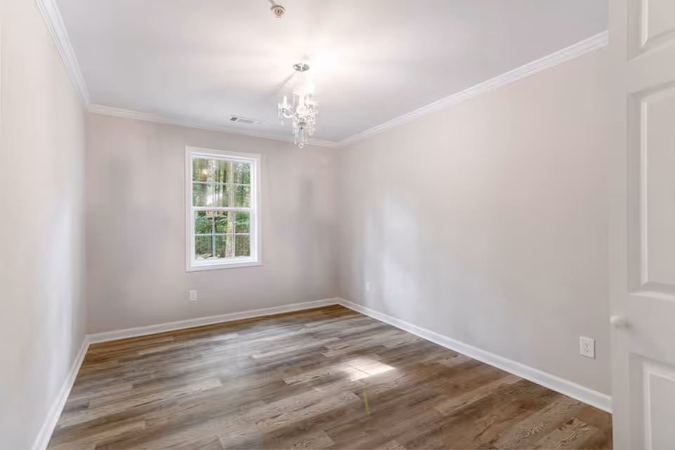 Empty small bedroom with a window, chandelier, neutral walls, and wood-look flooring.