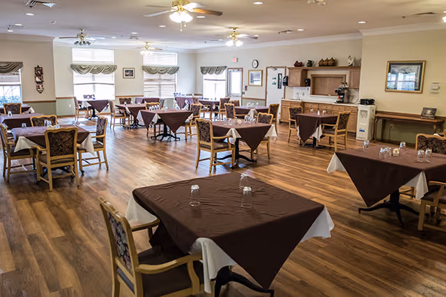 A spacious dining room with multiple tables covered with brown tablecloths and white underlays, each set with glasses. The room has wooden flooring, ceiling fans with lights, large windows with valances, and various framed pictures and decorations on the walls.