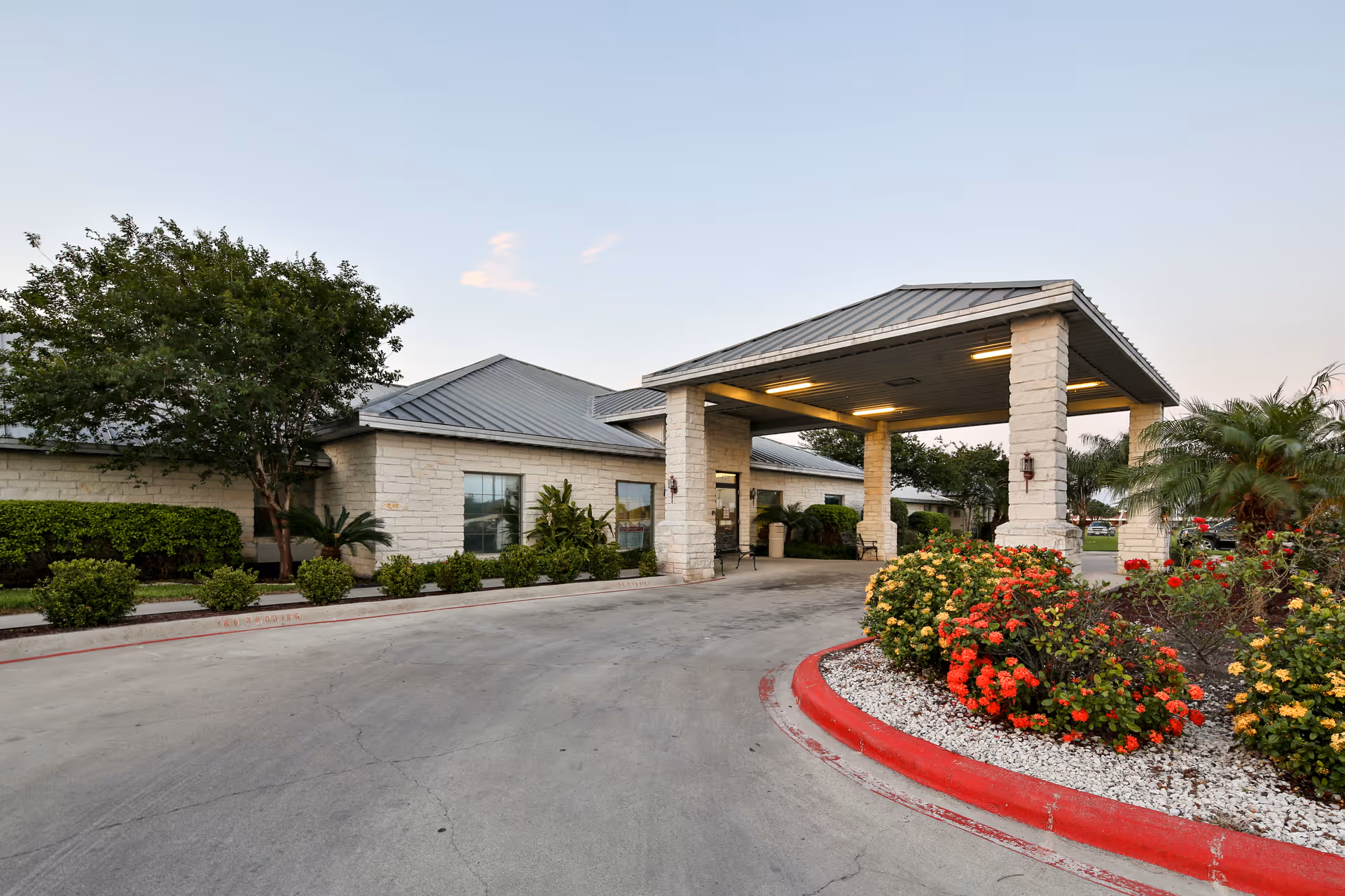 Exterior view of Weslaco Nursing and Rehabilitation Center showing the entrance with a covered drop-off area supported by stone pillars, surrounded by landscaped bushes and flowering plants under a clear sky.