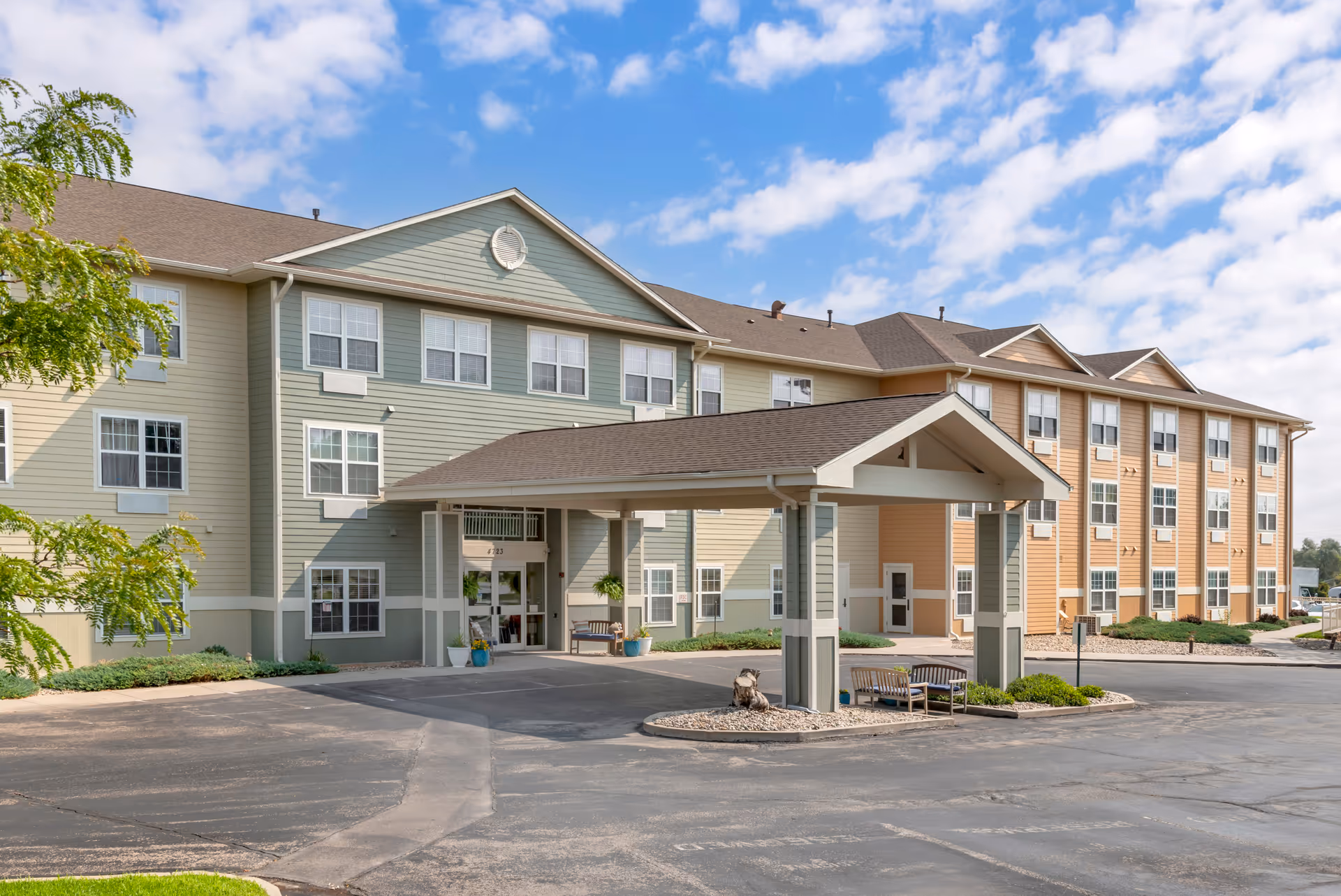 Front entrance of a multi-story senior living facility with a covered porte-cochere, benches, and parking area under a partly cloudy sky.