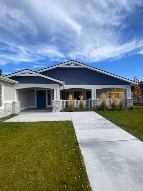 Single-story memory care building with a blue gabled facade, covered porch, white trim, a concrete walkway and lawn under a partly cloudy sky.
