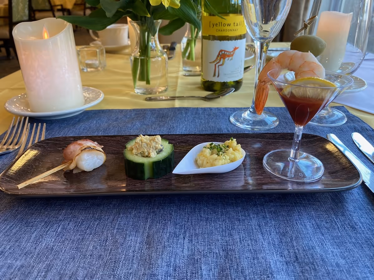 A rectangular wooden plate with four appetizers: a bacon-wrapped scallop on a skewer, a cucumber slice topped with a creamy mixture, a small serving of scrambled eggs garnished with chives in a white spoon, and a shrimp cocktail with a lemon slice in a small glass. The plate is on a blue placemat with a yellow tablecloth underneath. In the background, there is a lit candle, a bottle of Yellow Tail Chardonnay wine, a wine glass, and a vase with green leaves.