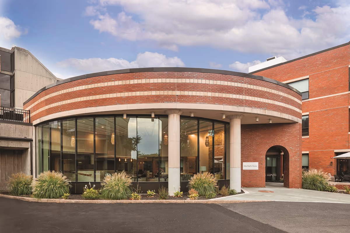 Exterior view of a modern assisted living facility building with a curved brick facade, large glass windows, and two white columns at the entrance. There are plants and shrubs along the building, and the sky is partly cloudy.