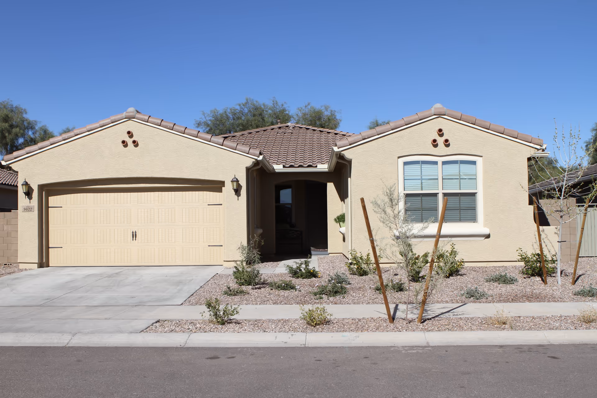 Front exterior view of a single-story beige house with a tiled roof, a two-car garage, a small landscaped front yard with desert plants, and a clear blue sky.