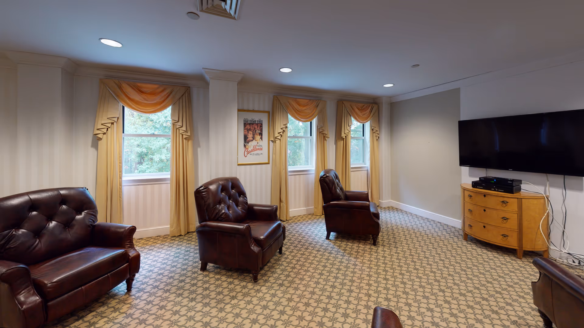 A cozy living room with three dark brown leather armchairs and one matching loveseat arranged on a patterned carpet. The room has three windows with yellow drapes and valances, a wall-mounted flat-screen TV above a wooden dresser, and a framed Casablanca movie poster on the wall between the windows.