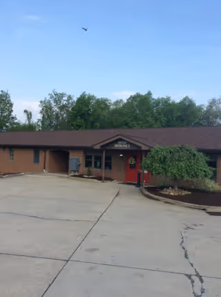 Exterior view of a single-story brick building with a brown roof and a red door labeled 'Main Entrance'. The building is surrounded by a paved driveway and some landscaping with trees and bushes. The sky is clear with a bird flying above.