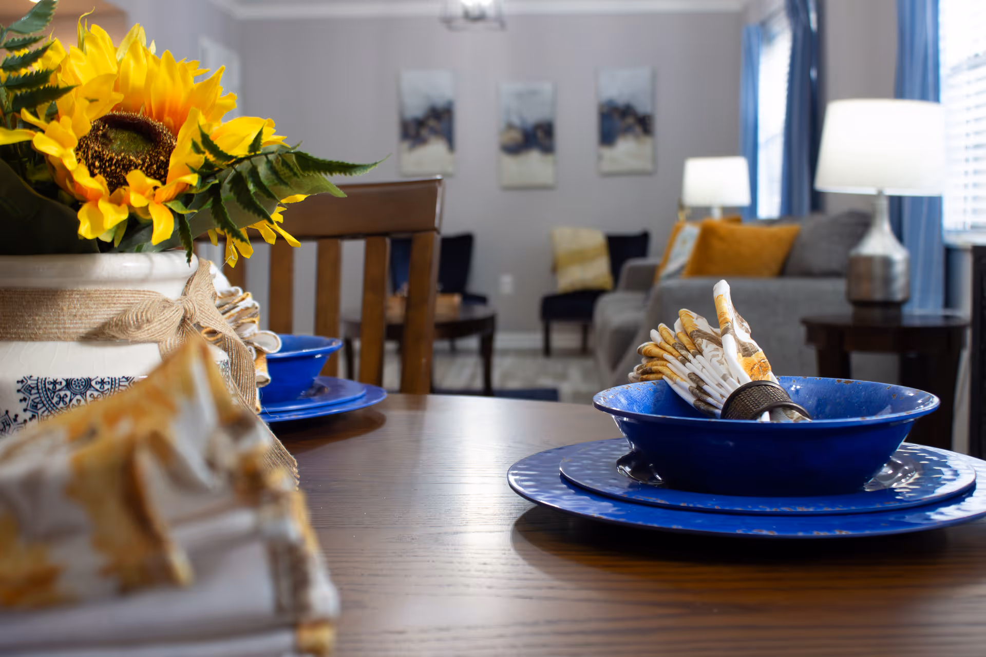 Close-up of a dining table set with blue plates and napkins and a sunflower centerpiece, with a living room visible in the background.