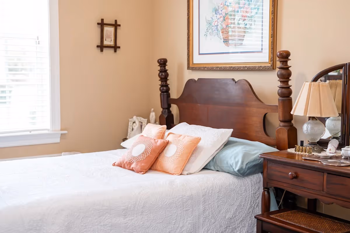 A cozy bedroom with a wooden bed frame featuring turned posts, a white quilted bedspread, and four pillows in white, peach, and light blue. Next to the bed is a wooden nightstand with a lamp, decorative items, and a mirror. A framed floral picture hangs on the beige wall above the bed, and a window with white blinds is on the left side.