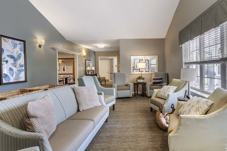 A bright and cozy living room area in a senior living facility with multiple comfortable armchairs and a sofa arranged around small tables with lamps. Large windows on the right side let in natural light, and framed artwork decorates the walls. The room has a neutral color palette with soft green and beige tones.