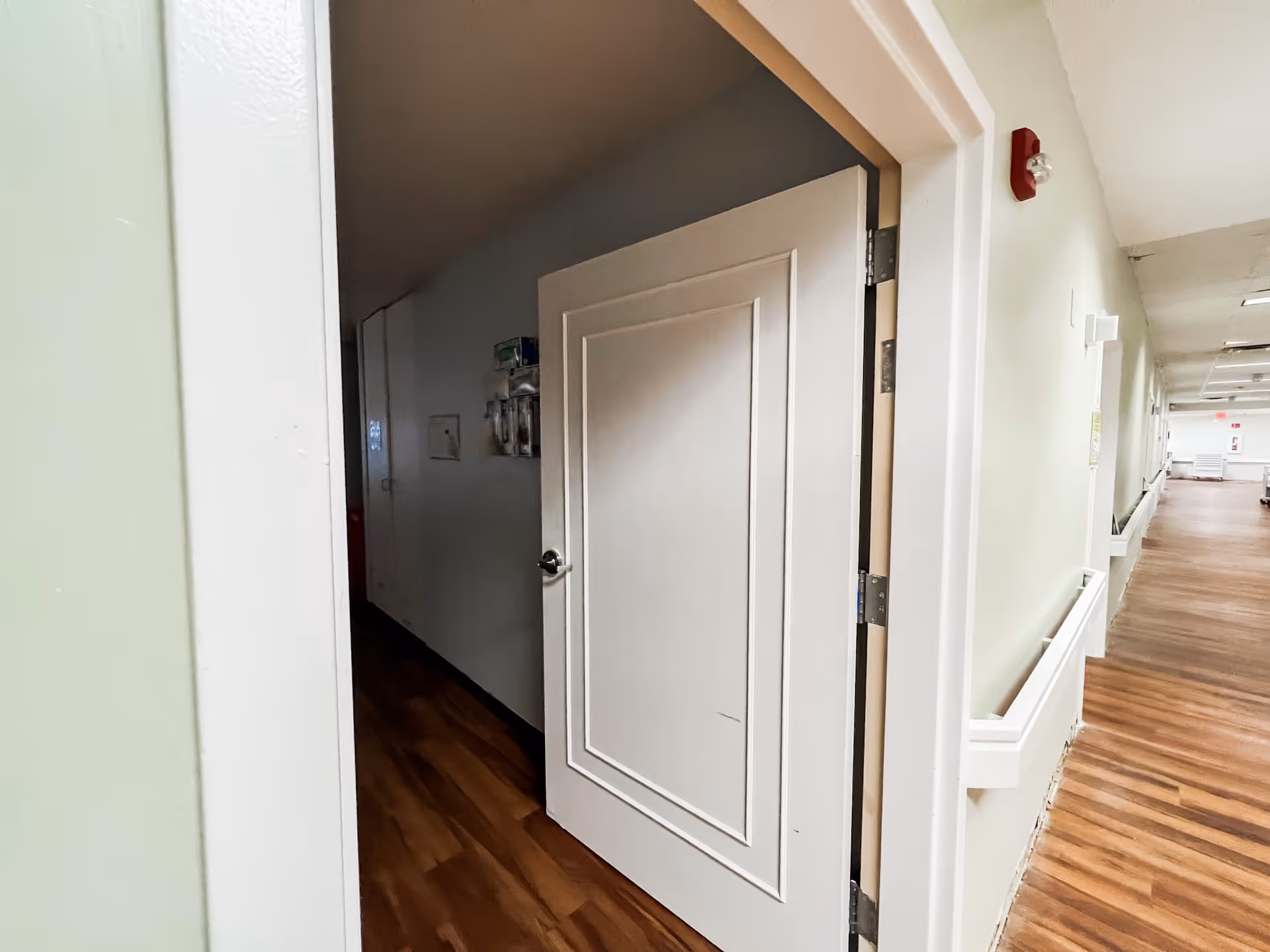 A hallway in a senior living facility with wood-patterned flooring and light-colored walls. A white door is partially open, revealing a darker interior space. The hallway has handrails on the walls and ceiling lights, with a fire alarm and emergency equipment visible.