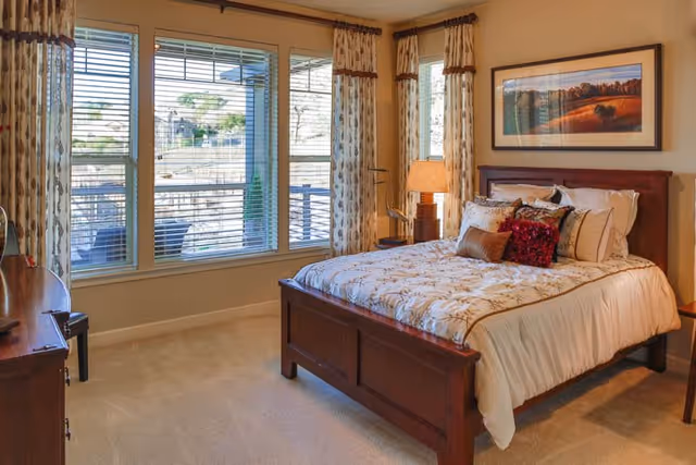A cozy bedroom with a wooden bed frame and a neatly made bed featuring floral-patterned bedding and decorative pillows. The room has large windows with blinds and patterned curtains, allowing natural light to fill the space. A wooden dresser and a bedside table with a lamp are also visible, along with a framed landscape painting on the wall.