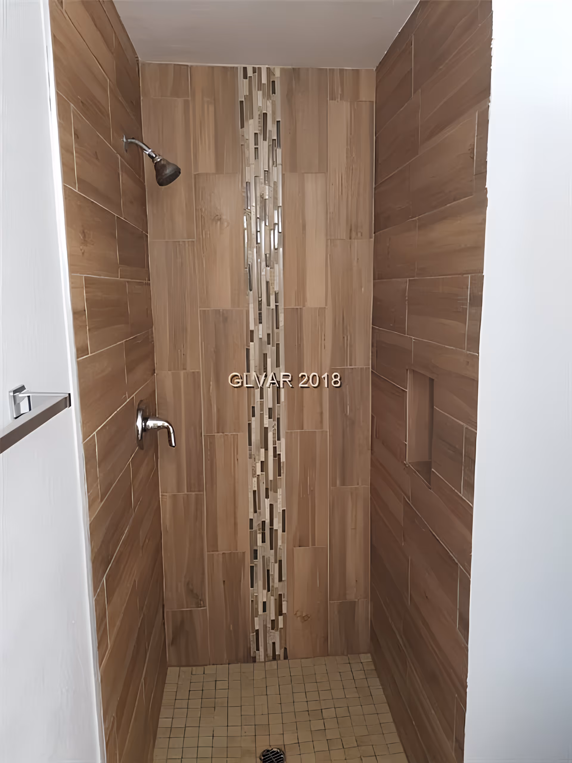 A shower area with brown wood-like tiles on the walls and a vertical strip of decorative mosaic tiles in the center. The floor has small square beige tiles. There is a silver showerhead and a matching faucet on the left wall, and a recessed shelf on the right wall.