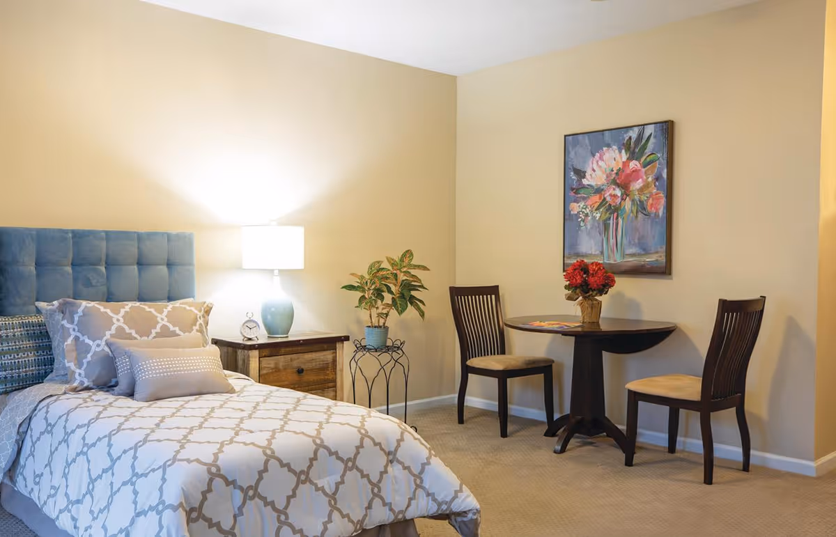 A cozy bedroom with a bed featuring a blue tufted headboard and patterned bedding. Next to the bed is a wooden nightstand with a blue lamp and a small clock. A potted plant sits on a decorative metal stand. In the corner, there is a small round wooden table with two chairs, a vase with red flowers, and a colorful floral painting on the wall.