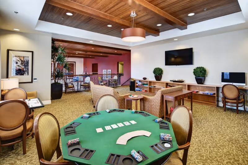 A spacious common area in a senior living facility featuring a green poker table with playing cards and chips, surrounded by chairs. In the background, there are two beige sofas facing a wall-mounted TV, a wooden shelf with board games, and a computer desk with a chair. The room has a carpeted floor, wooden ceiling beams, and warm lighting.