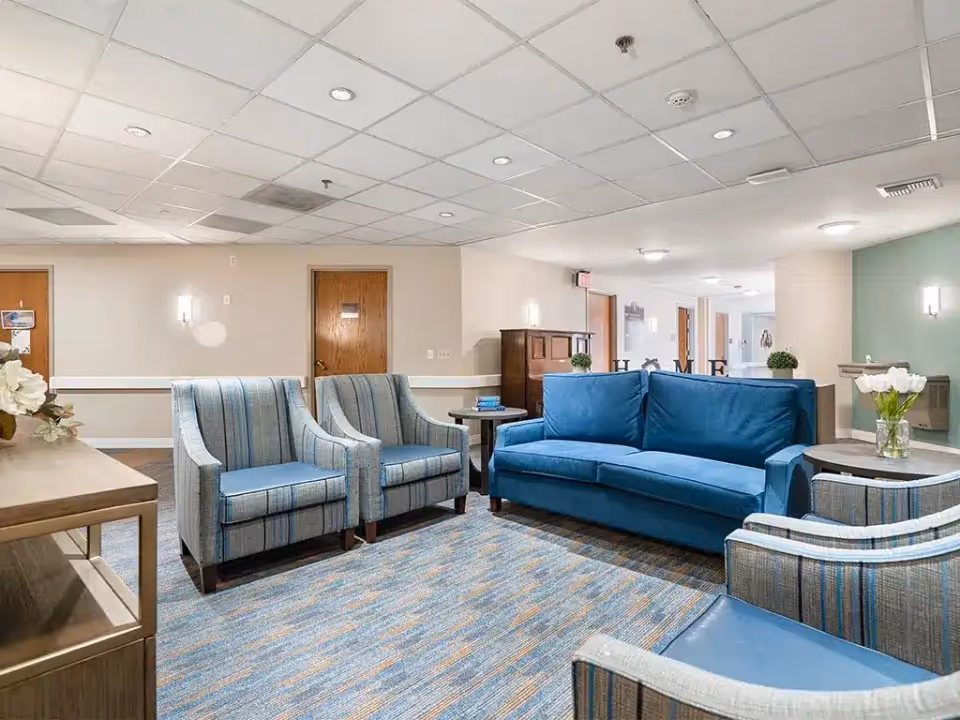 A cozy seating area in a senior living facility with a blue sofa, two blue striped armchairs, a wooden side table with a vase of white flowers, and a patterned carpet. The background shows a hallway with wooden doors and soft lighting fixtures on the walls.