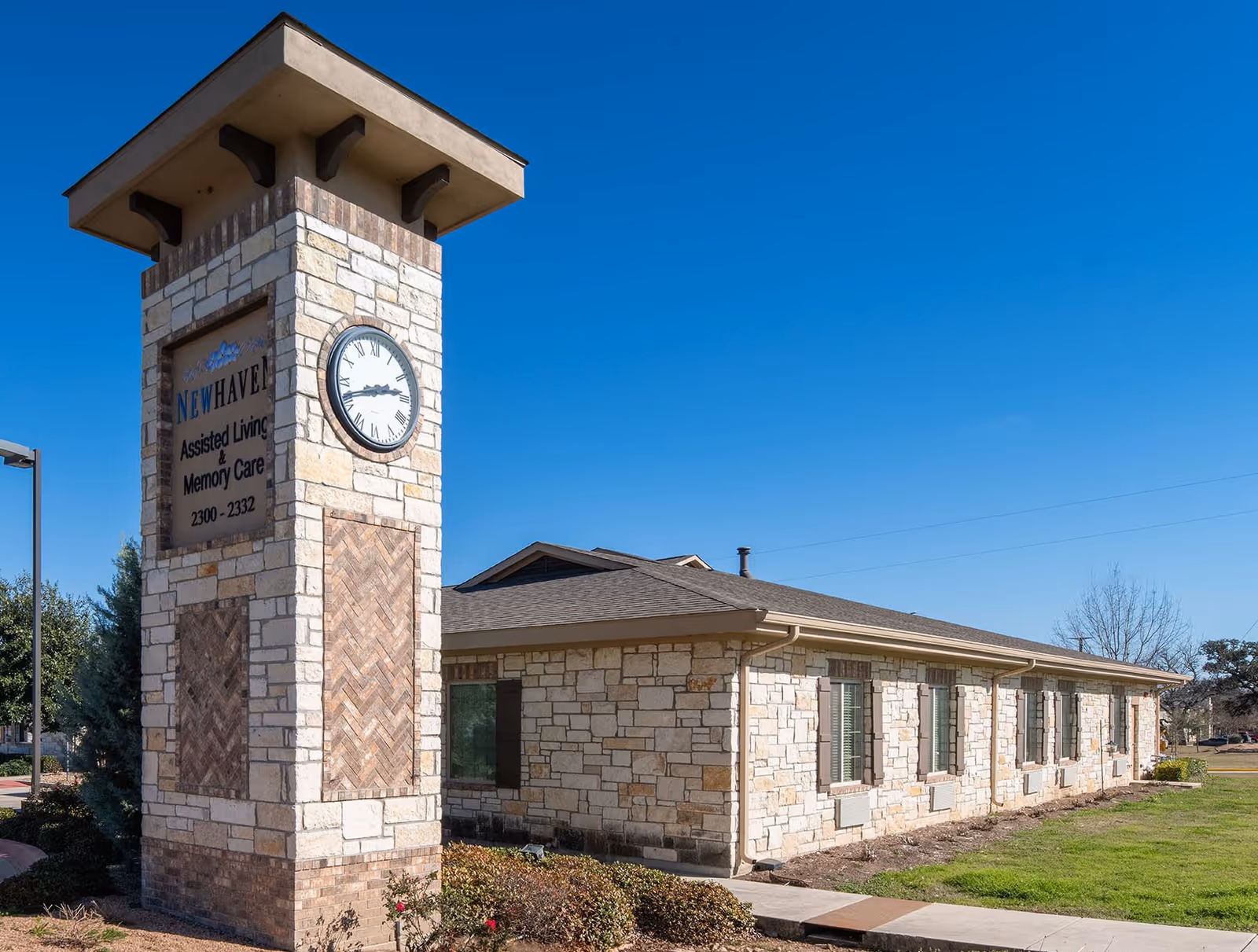 Exterior view of New Haven Assisted Living and Memory Care of Schertz building with a stone clock tower displaying the time. The building is made of light-colored stone with several windows and a well-maintained lawn under a clear blue sky.