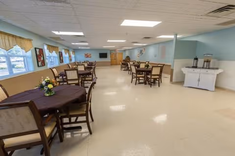 A spacious dining room with multiple tables and chairs arranged neatly. The tables are covered with dark tablecloths and decorated with small flower arrangements. Large windows with yellow valances allow natural light to fill the room. The walls are painted light blue and beige, and there is a white cabinet with beverage dispensers on the right side. The ceiling has recessed lighting panels.