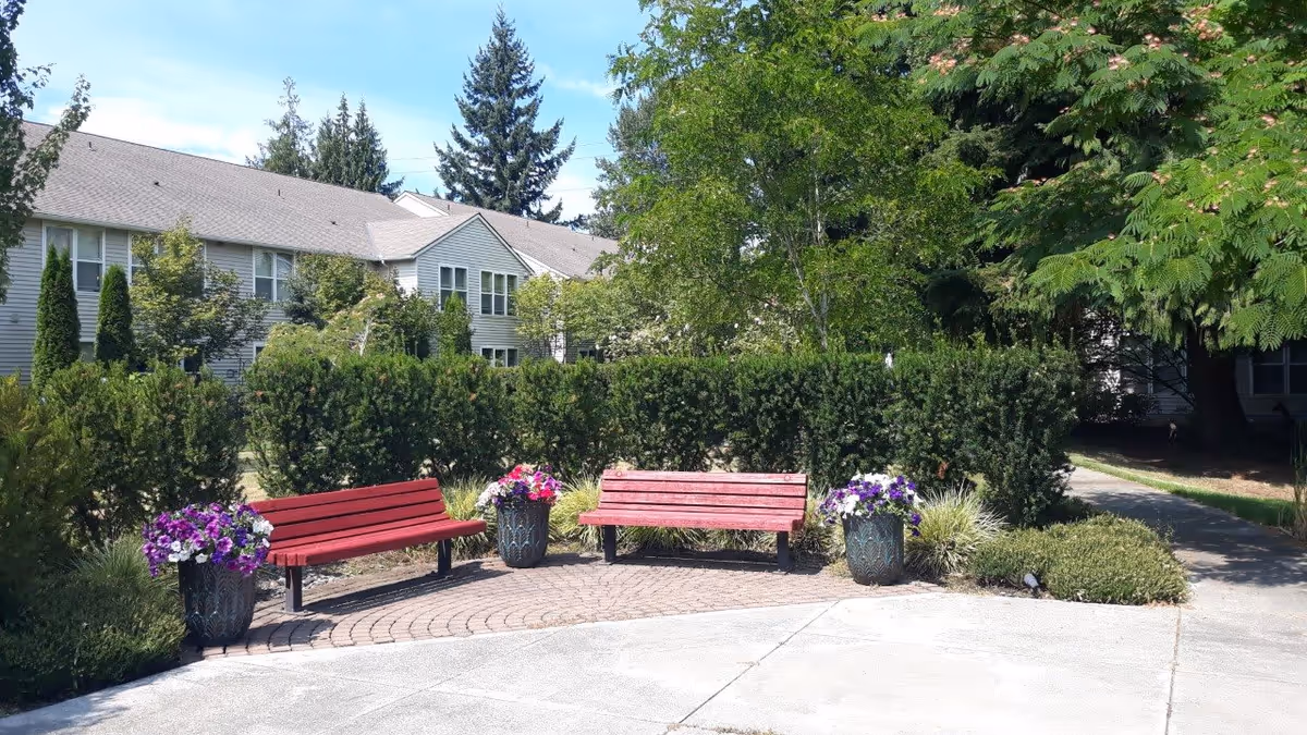 Outdoor seating area with two red benches and two large flower pots filled with purple and white flowers, surrounded by green bushes and trees, with a building visible in the background under a partly cloudy sky.