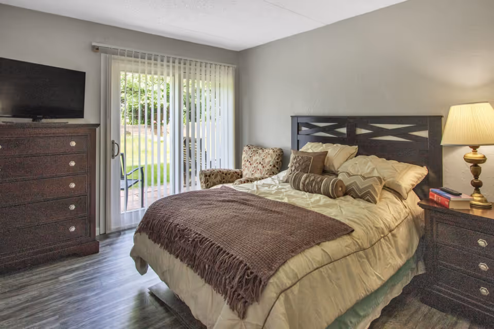 A cozy bedroom with a neatly made bed featuring multiple pillows and a brown throw blanket. There is a wooden nightstand with a lamp and books on the right side of the bed. To the left, there is a wooden dresser with a flat-screen TV on top. A patterned armchair is placed near sliding glass doors with vertical blinds, leading to an outdoor patio with chairs and greenery visible outside.