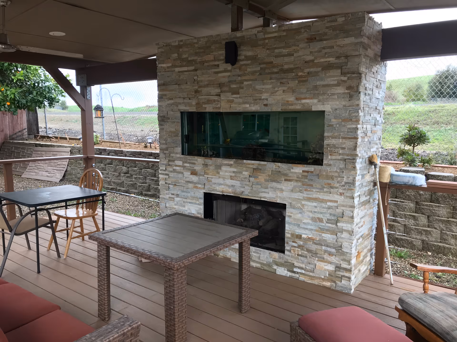 Covered outdoor patio area with a stone fireplace that has a built-in aquarium above it. The patio has wooden flooring, a small table with chairs, and some seating furniture. There is a fence and greenery visible in the background.