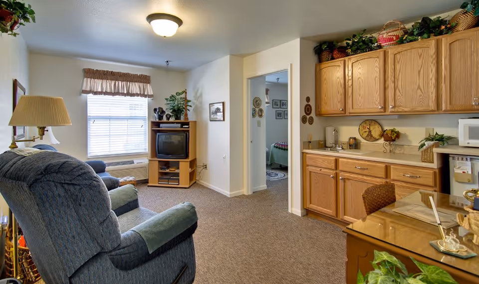 A cozy living area in a senior living facility with a blue recliner chair, a wooden TV stand holding an older model television, a window with blinds and a valance, and a small kitchenette with wooden cabinets, a sink, microwave, and a glass-top table with a wicker chair. There is a doorway leading to a bedroom with a bed and framed pictures on the wall.