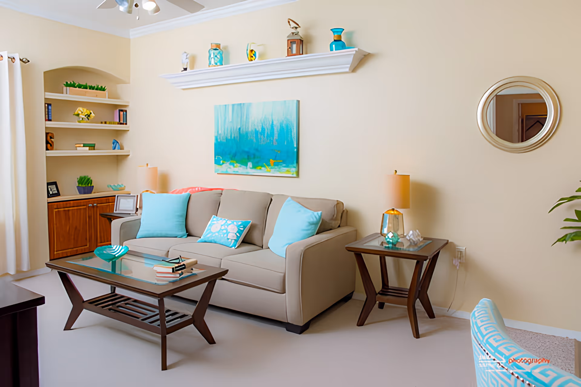 Sunny living room with a beige sofa topped with teal pillows, a glass coffee table, side tables with lamps, wall art and built-in shelving.