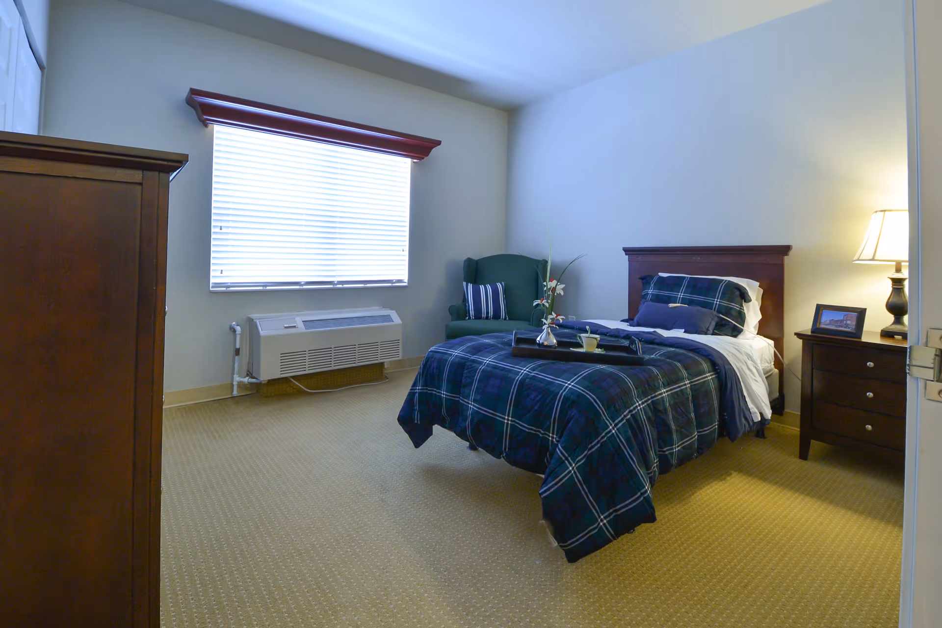 A neatly arranged bedroom with a single bed covered in a dark plaid comforter and pillows. A wooden nightstand with a lamp and a framed picture is beside the bed. There is a green armchair with a striped pillow in the corner near a window with closed blinds. A wooden dresser is partially visible on the left side of the image. The room has beige carpet and light-colored walls.