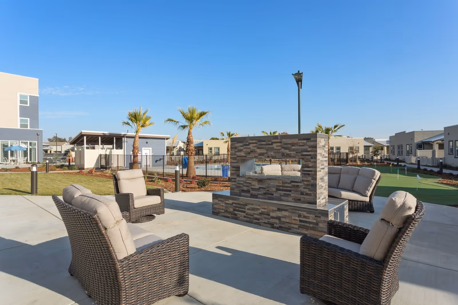 Outdoor seating area with four cushioned wicker chairs arranged around a modern rectangular stone fireplace. In the background, there are palm trees, a fenced swimming pool, a small putting green, and several single-story buildings under a clear blue sky.