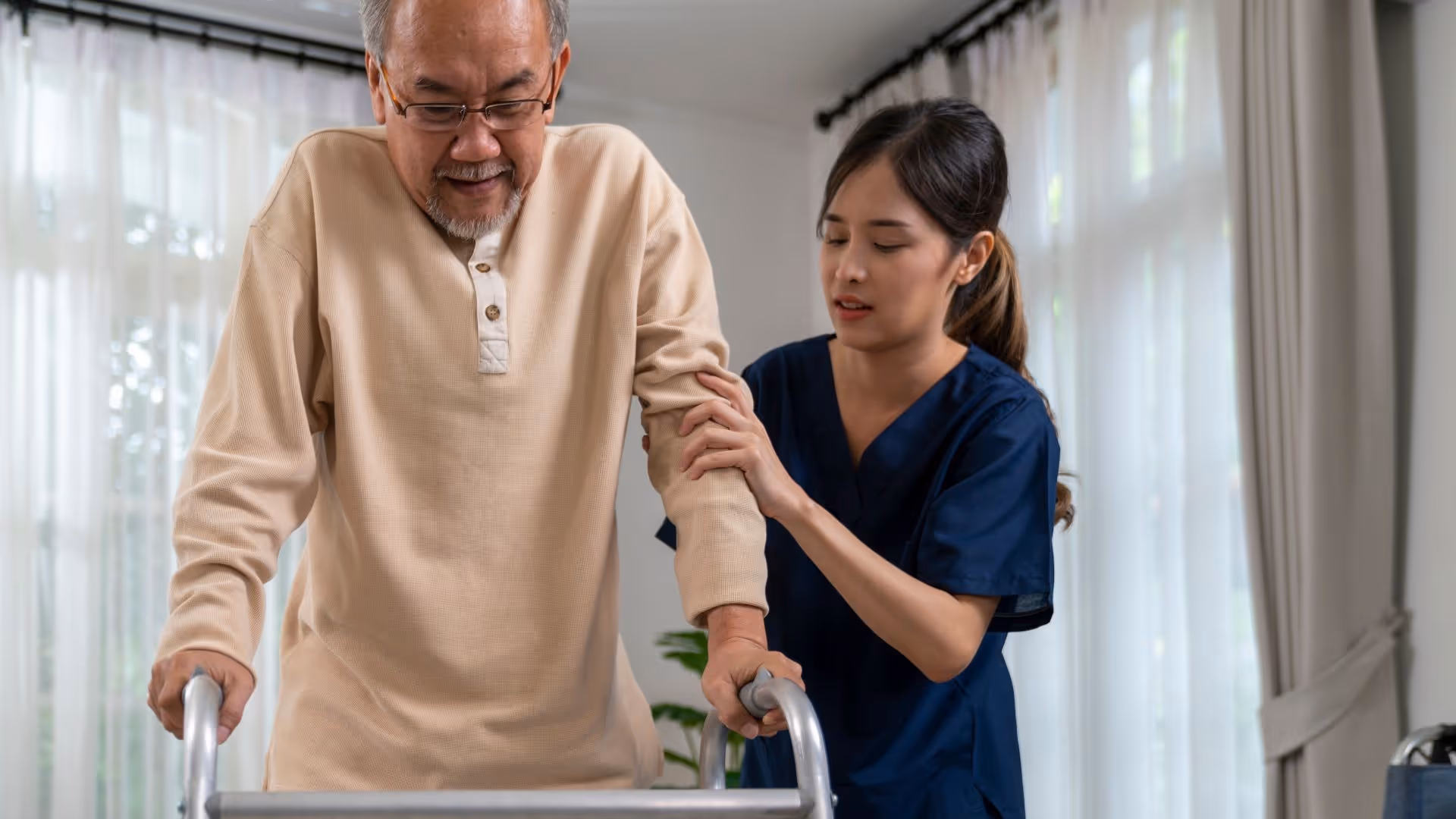 A caregiver in navy scrubs assists an elderly man using a walker inside a well-lit room with large windows and light curtains.