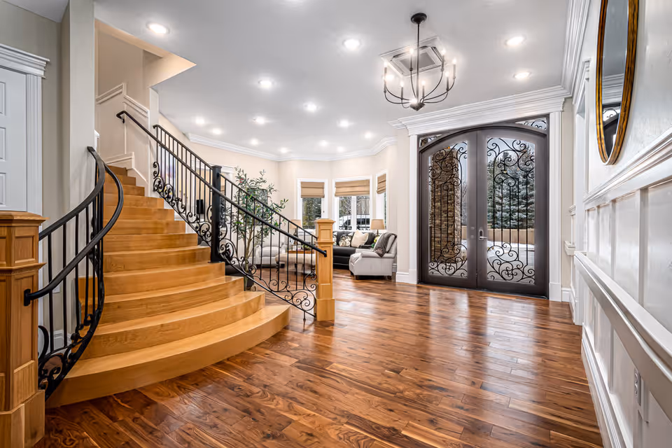 Spacious and elegant interior entryway featuring a curved wooden staircase with ornate black metal railings, polished hardwood floors, a large decorative double door with intricate wrought iron designs, a chandelier hanging from the ceiling, and a cozy seating area with a sofa and armchair near bay windows with blinds.